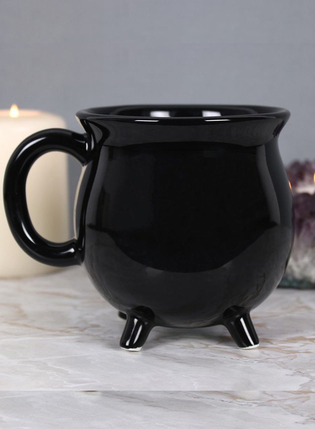Black Cauldron Mug – black glossy mug in the shape of a witch's cauldron with a round handle, wide opening and three small legs against a white background.